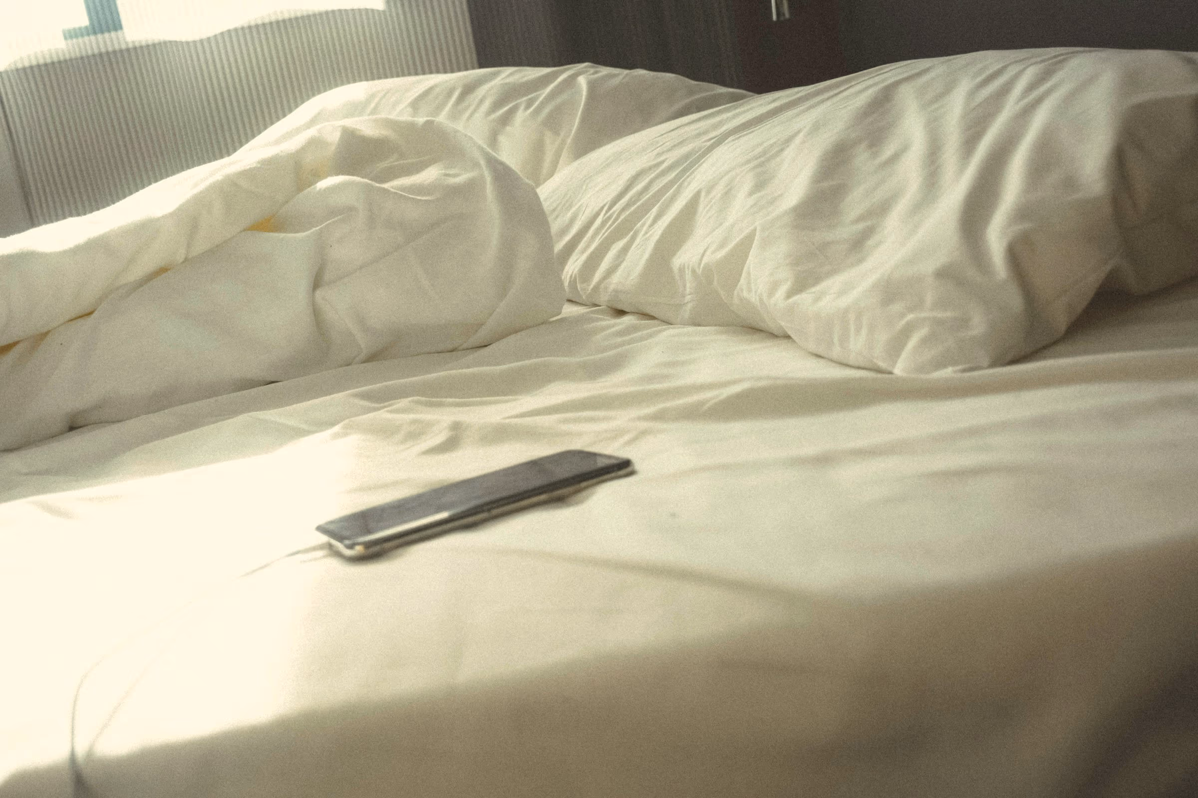 Smartphone écran éteint relié à son câble de charge, posé sur un drap blanc froissé à côté d’oreillers, lumière douce du matin