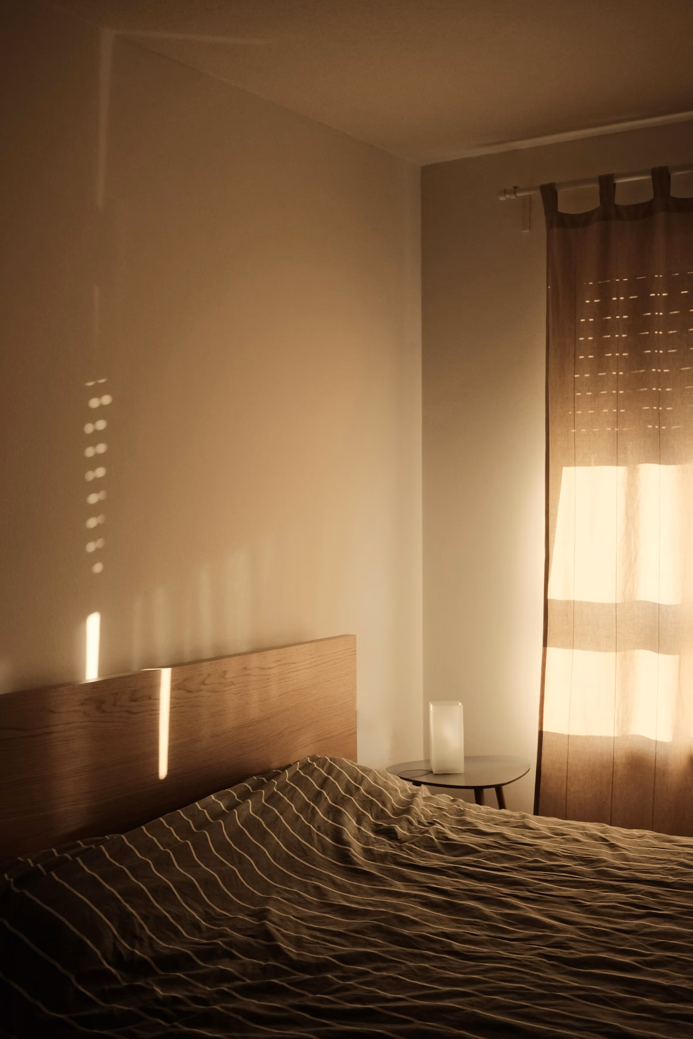 Chambre sobre : lit au couvre-lit rayé, tête de lit en bois et rideau filtrant une lumière chaude sur le mur