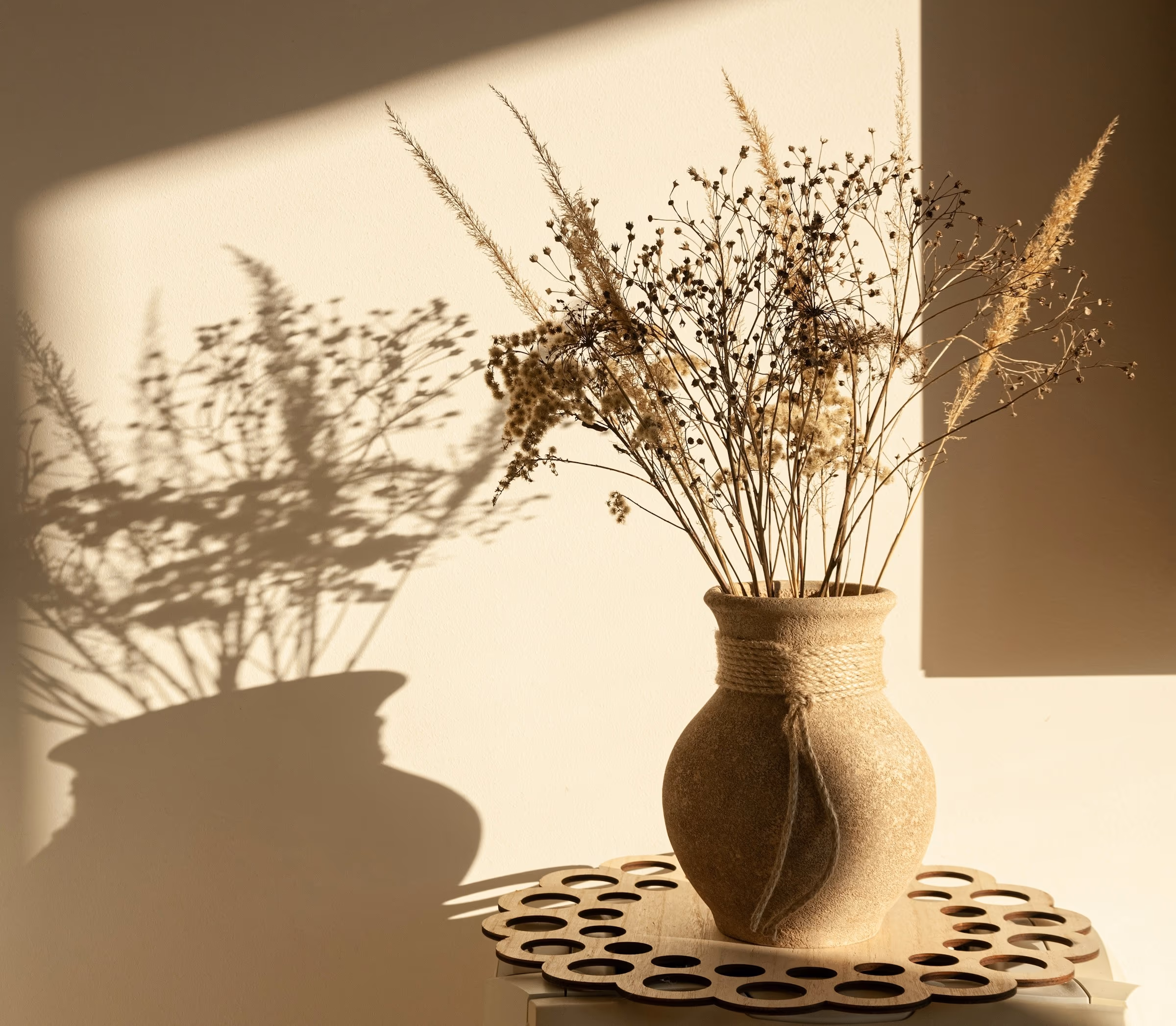 Un vase de céramique garni de fleurs séchées, posé dans la lumière chaude d’une fin de journée, l’ombre des tiges sur le mur