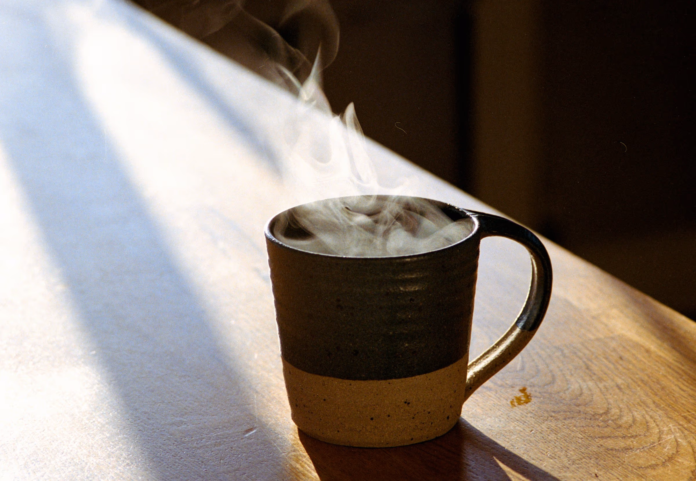 Tasse en grès bicolore d’où s’élève une vapeur, sur une table en bois traversée par un rai de soleil bas