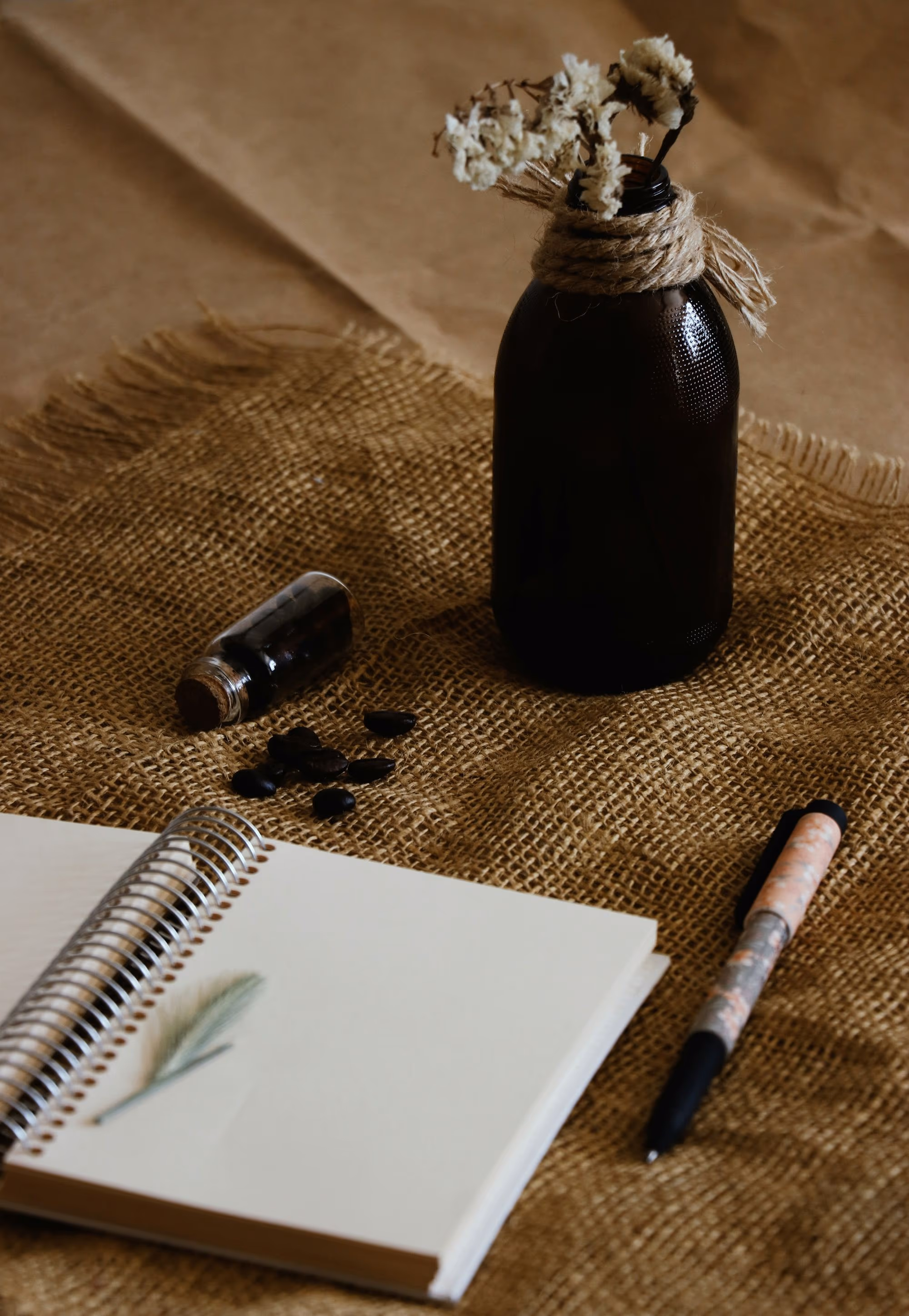 Carnet à spirale ouvert sur un brin d’herbe séché, avec un stylo, une fiole et une bouteille de fleurs séchées sur une toile de jute