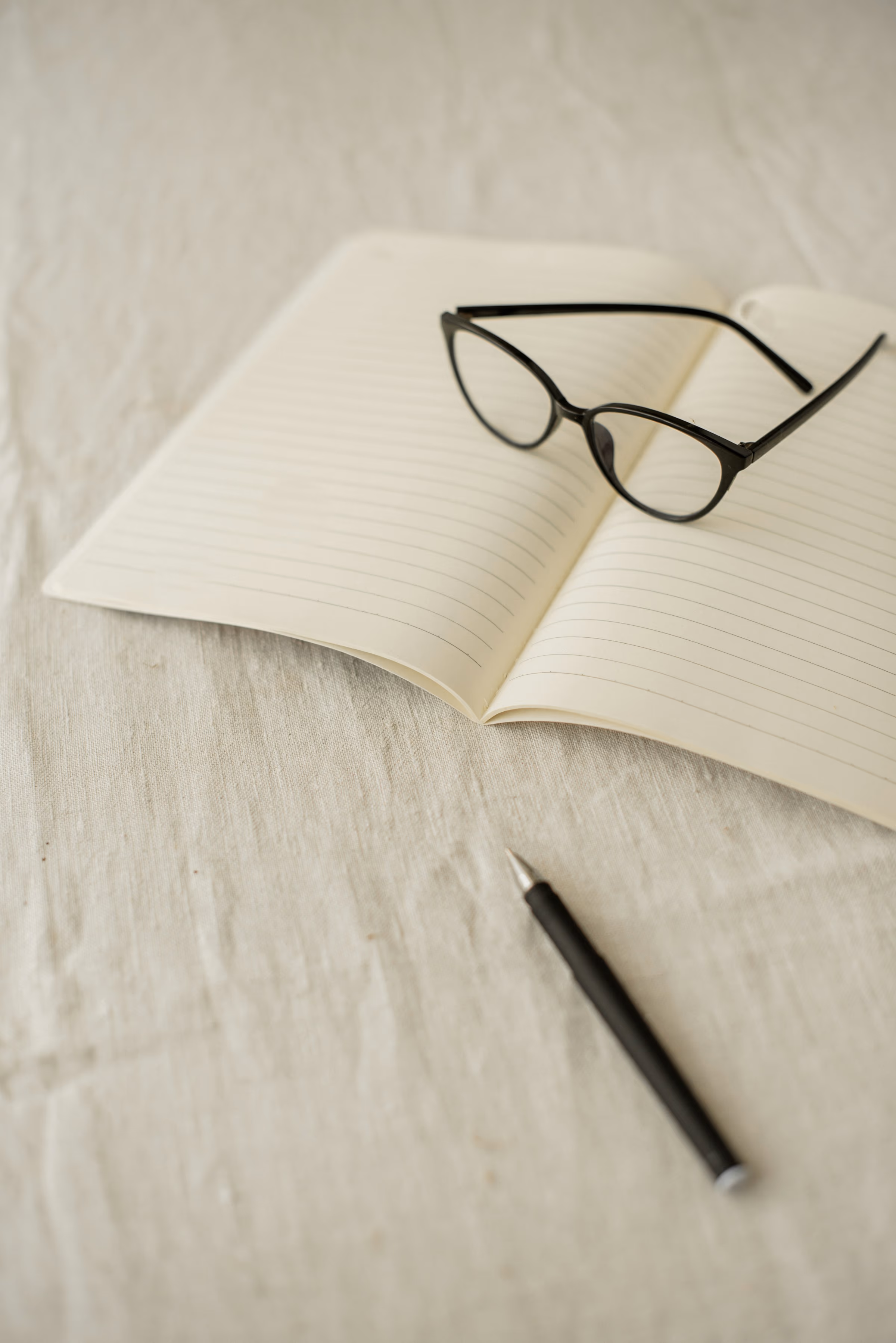 Cahier ouvert aux pages lignées, lunettes noires et stylo posés sur une nappe de lin naturel.