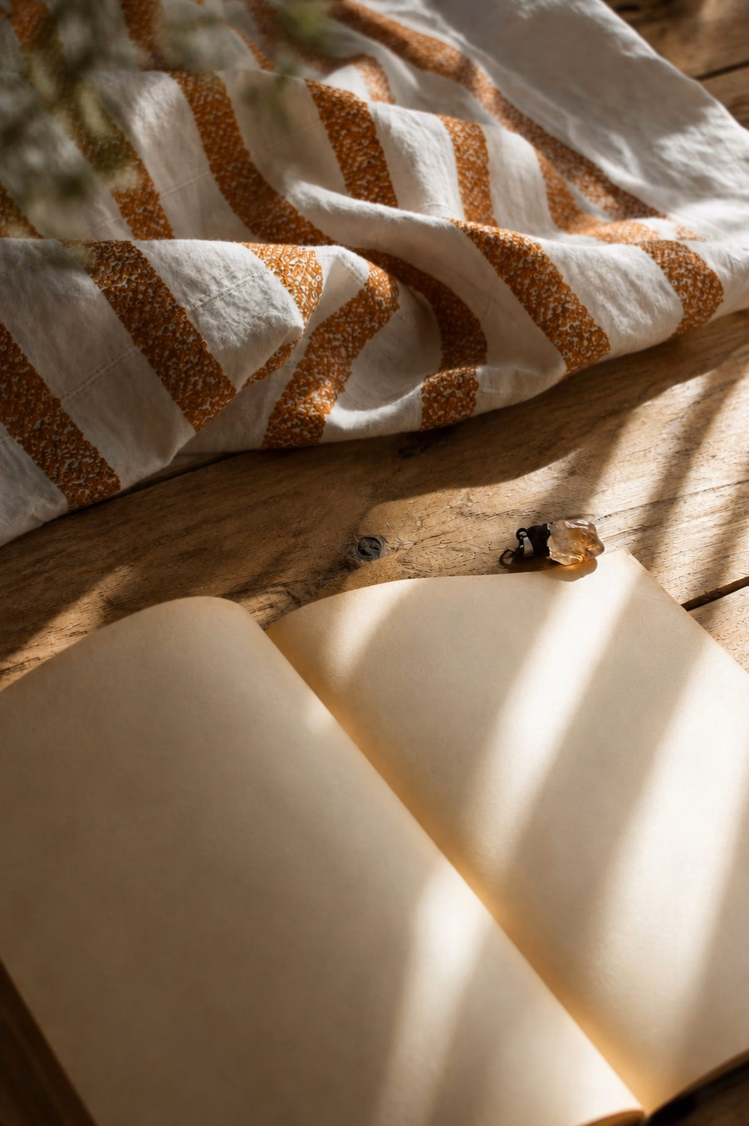 Carnet ouvert aux pages vierges sur une table en bois rustique, avec un petit cristal et un linge rayé ocre, sous une lumière naturelle.