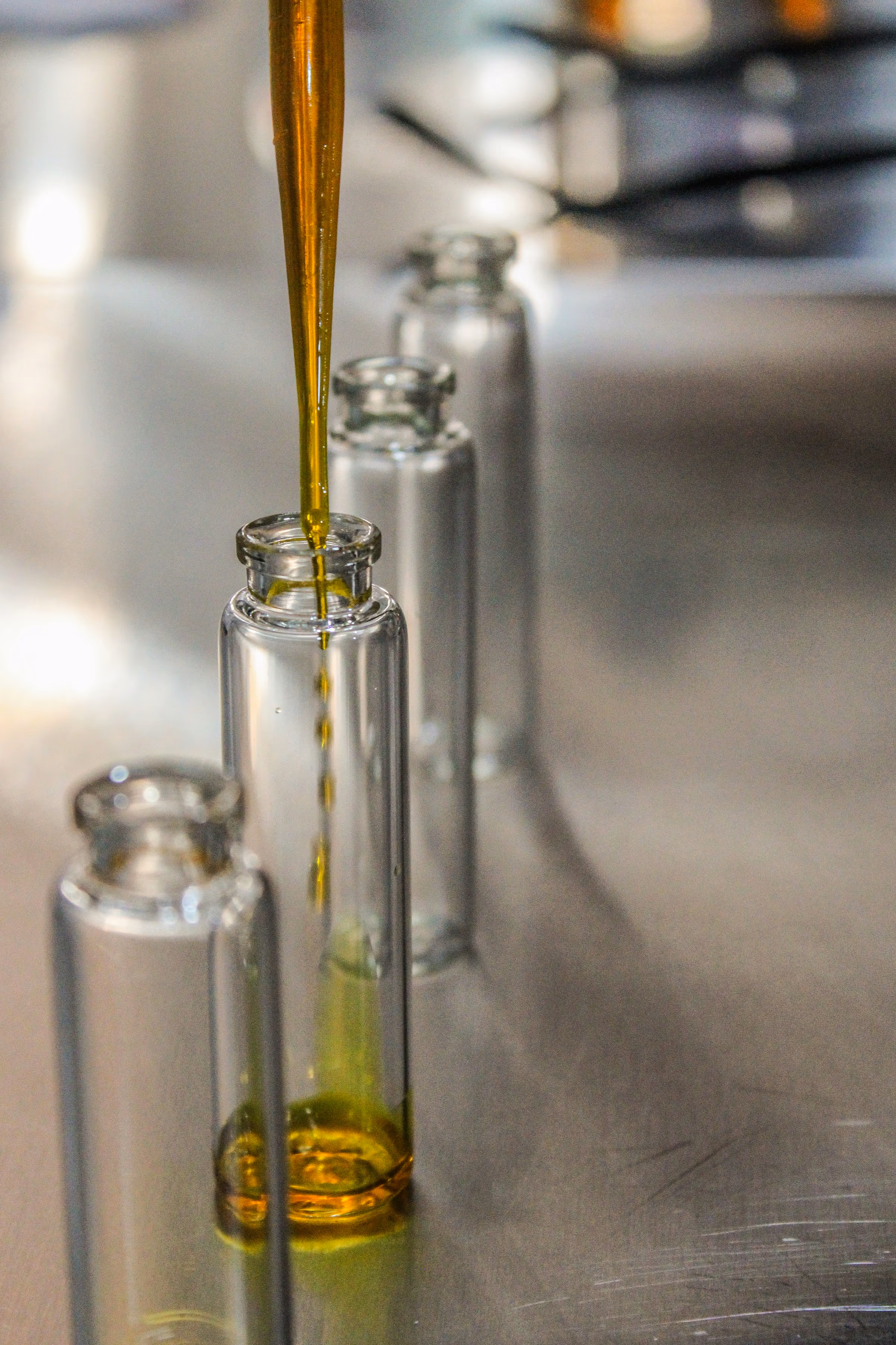 Filet d’huile dorée versé à la pipette dans des fioles en verre transparent sans inscription, sur un plan de travail métallique.