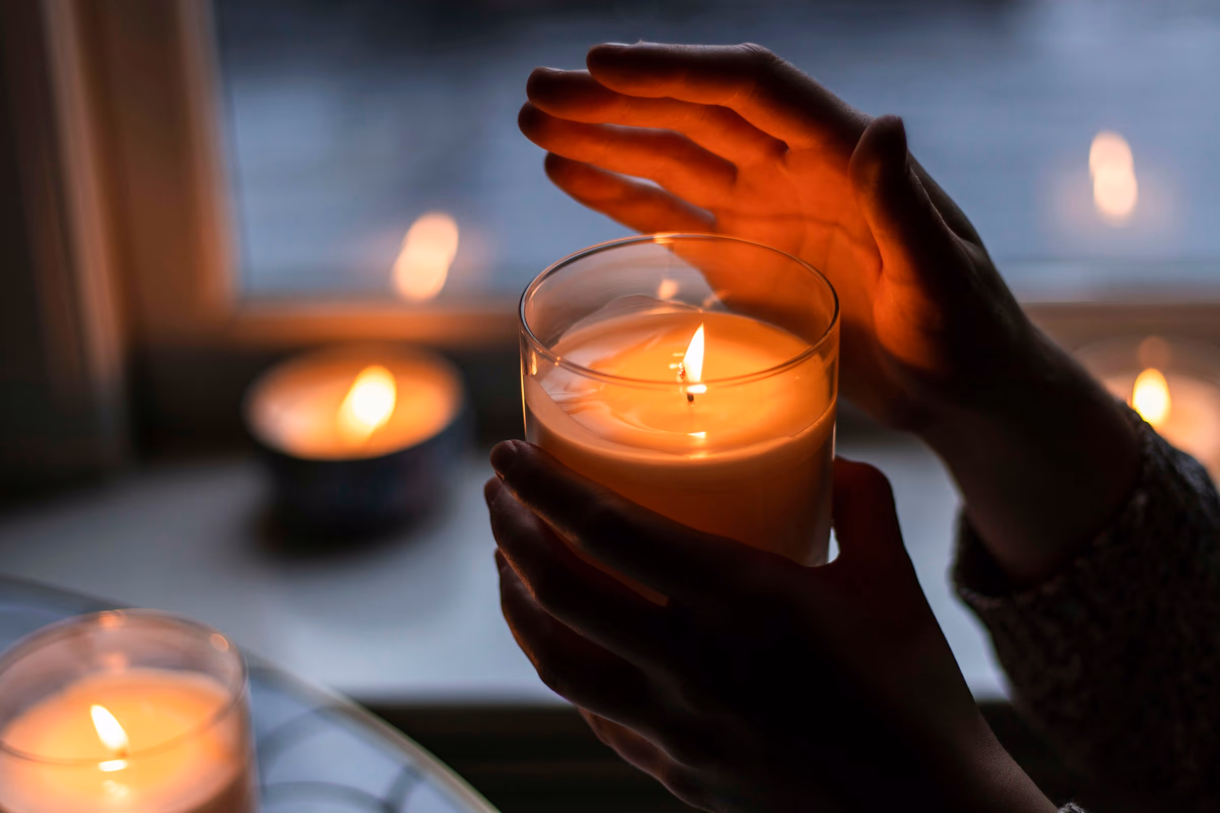 Des mains protègent la flamme d’une bougie, près d’une fenêtre, le soir