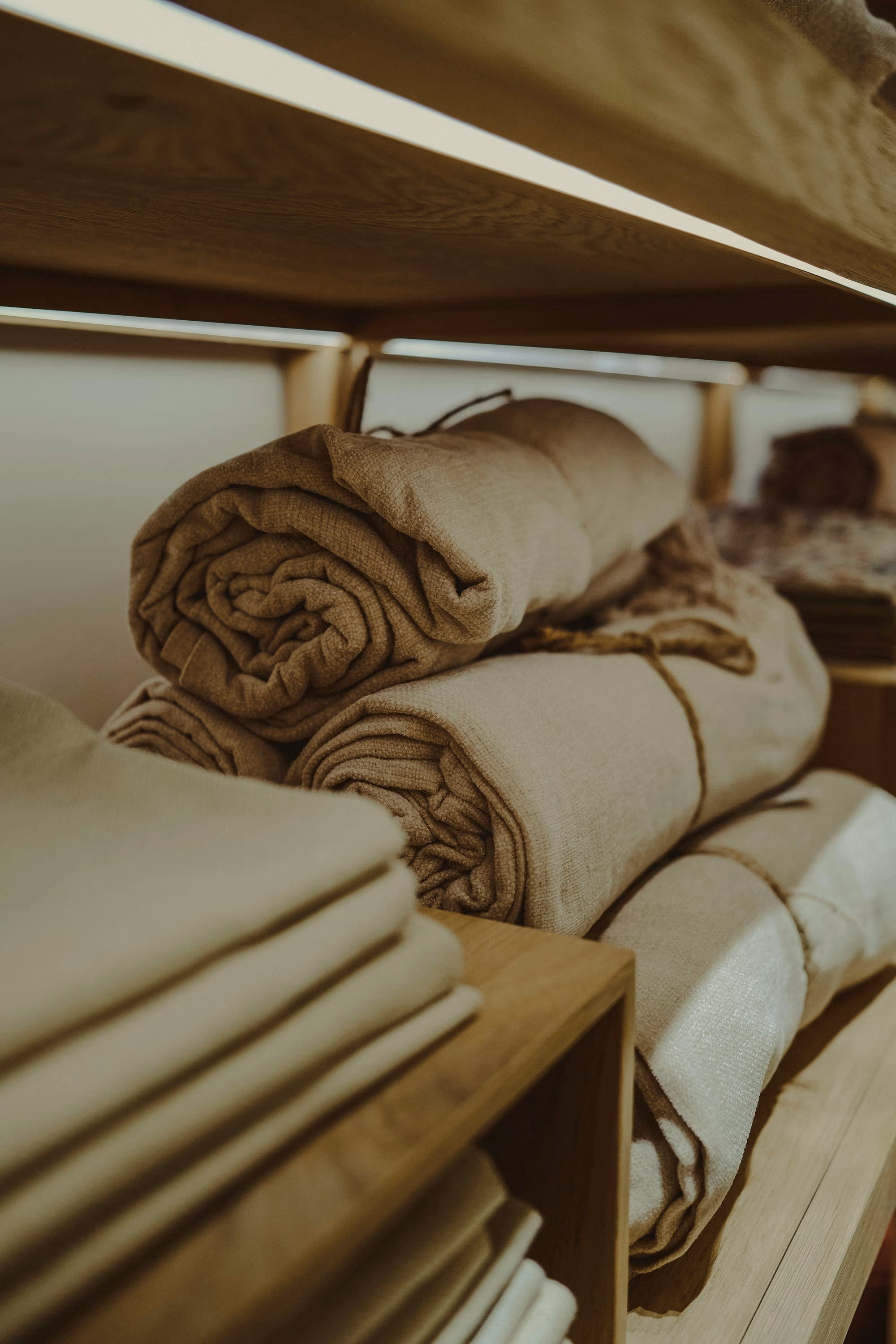 Linges de lin écru roulés et noués d’une ficelle sur une étagère en bois clair, à côté d’une pile de serviettes pliées.