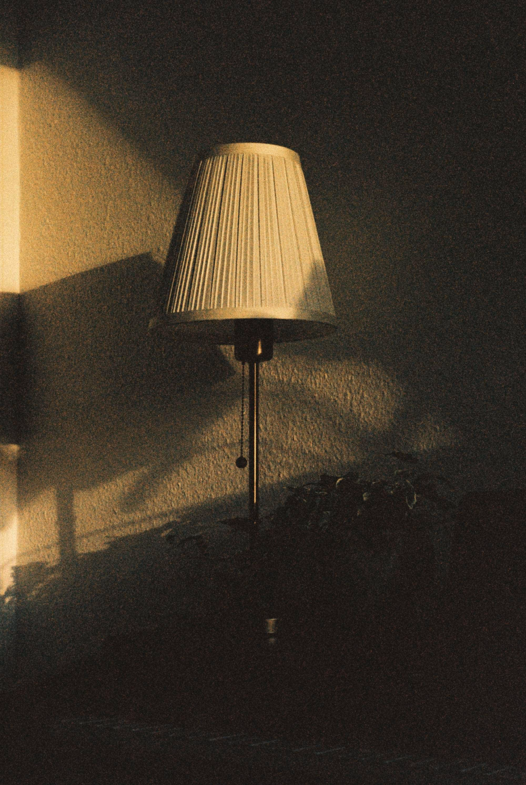 Lampe à abat-jour plissé baignée par un rayon de soleil oblique, son ombre portée sur un mur crépi
