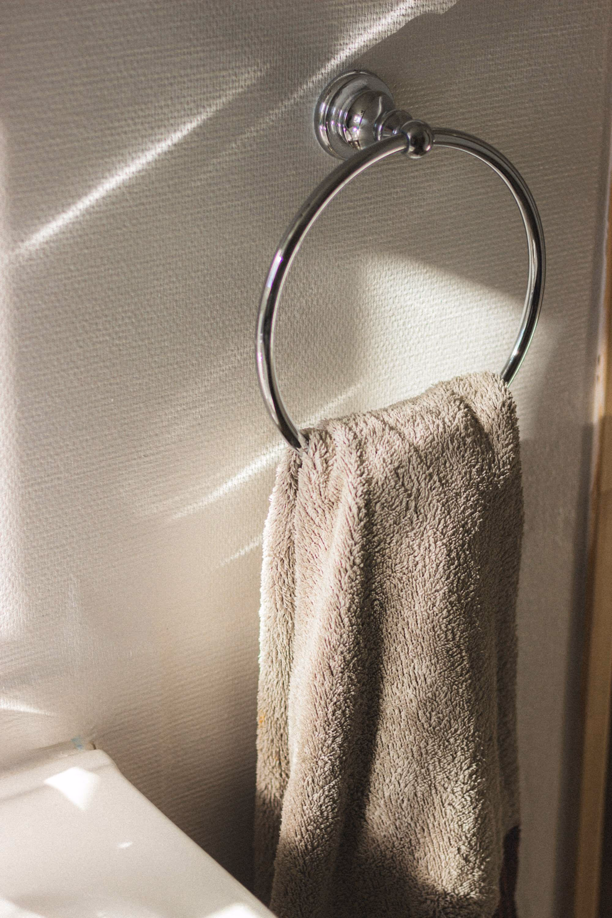 Serviette éponge taupe passée dans un anneau porte-serviette chromé, sur un mur crème strié de rais de soleil