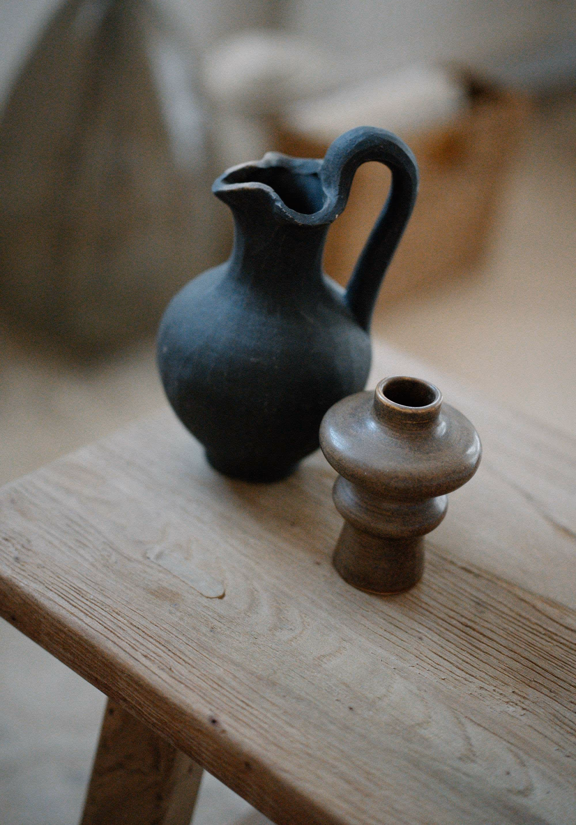 Pichet en terre sombre et petit vase en grès brun posés sur un banc en bois clair, dans la lumière douce d’un intérieur aux tons beiges.