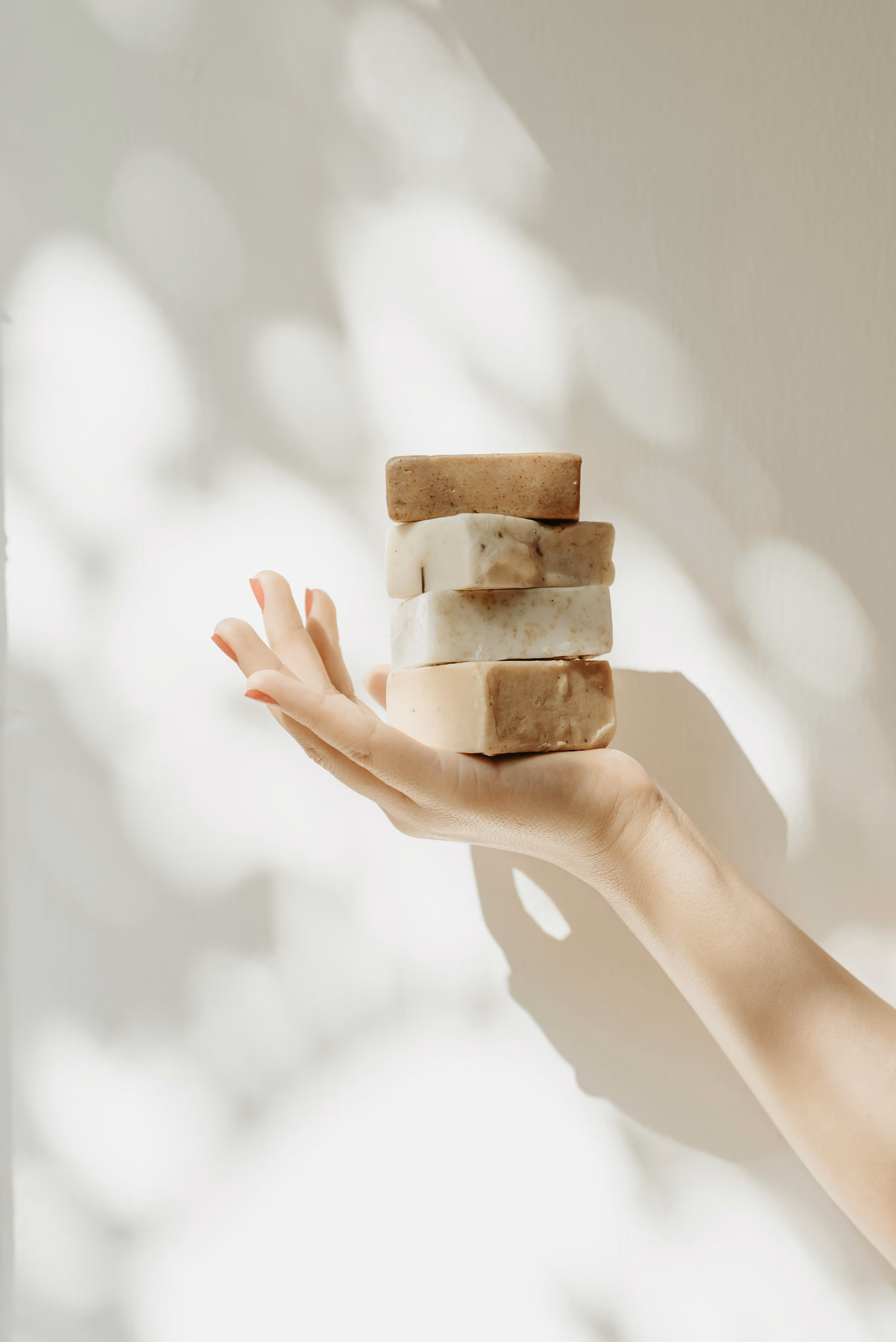 Main tendue présentant une pile de quatre savons artisanaux, devant un mur crème marqué d’ombres de feuillage