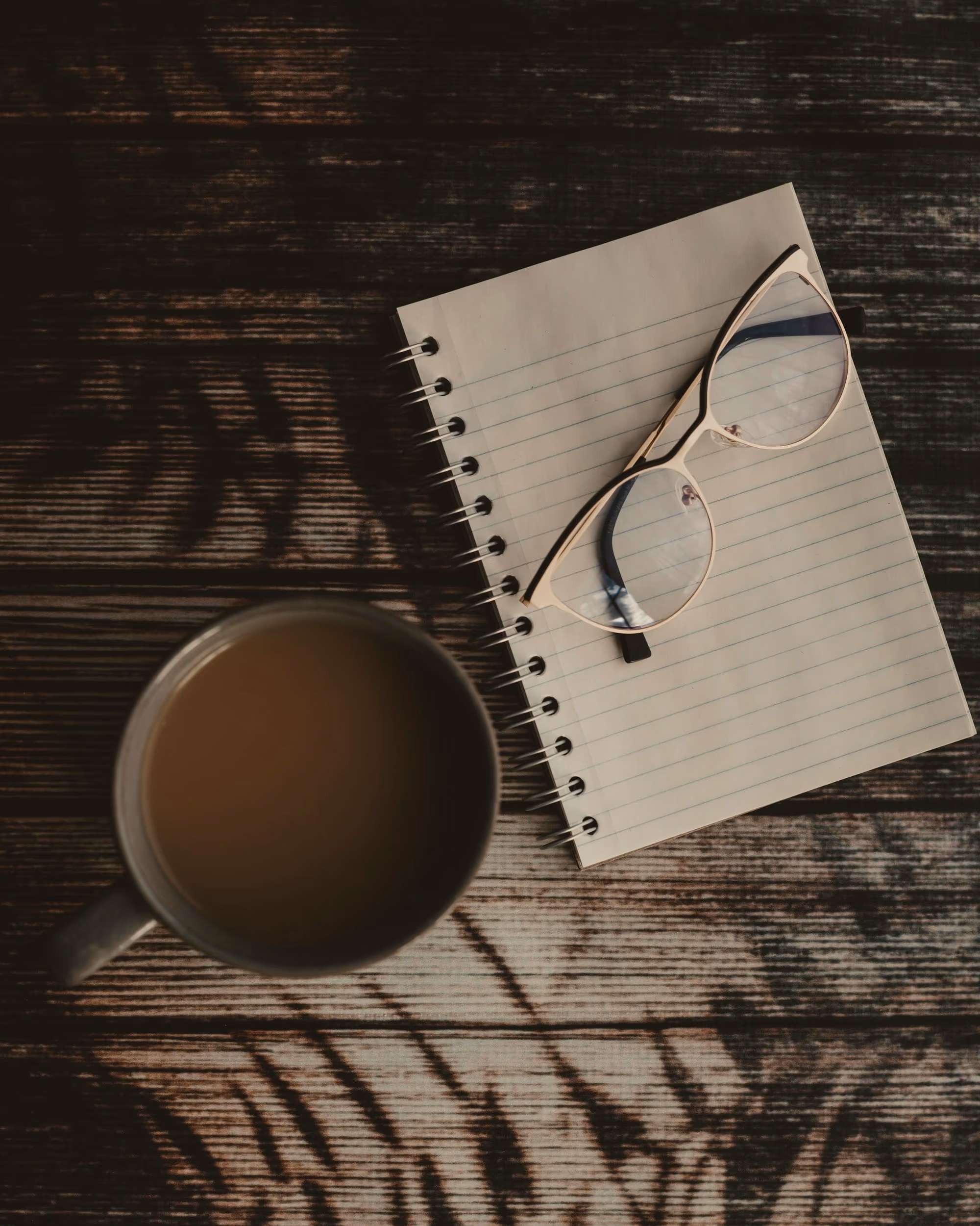 Vue plongeante : tasse de café au lait, carnet ligné vierge et lunettes posés sur une table en bois traversée d’ombres de feuillage