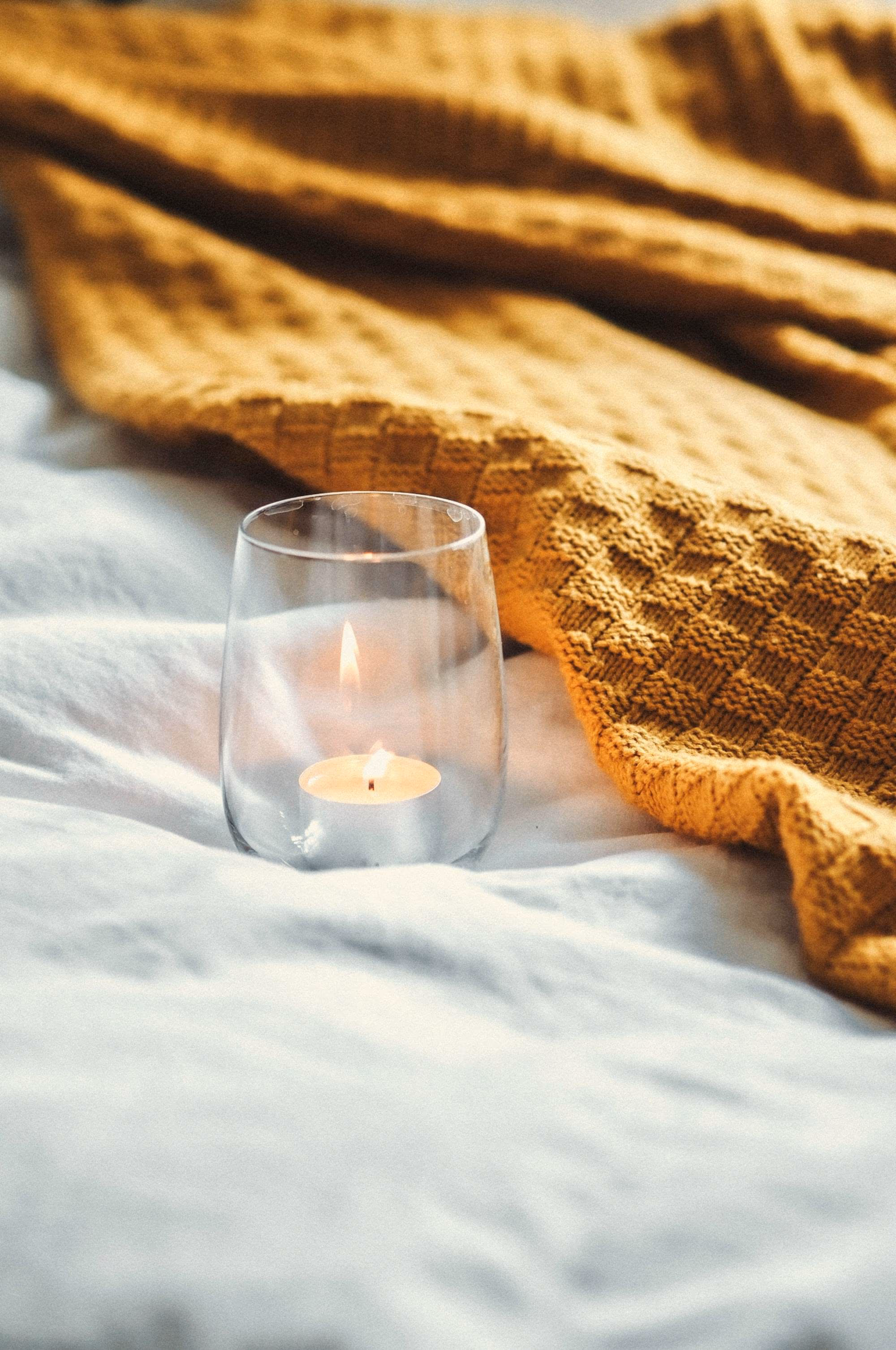 Photophore en verre et bougie allumée posés sur un drap blanc, près d’un plaid gaufré moutarde.