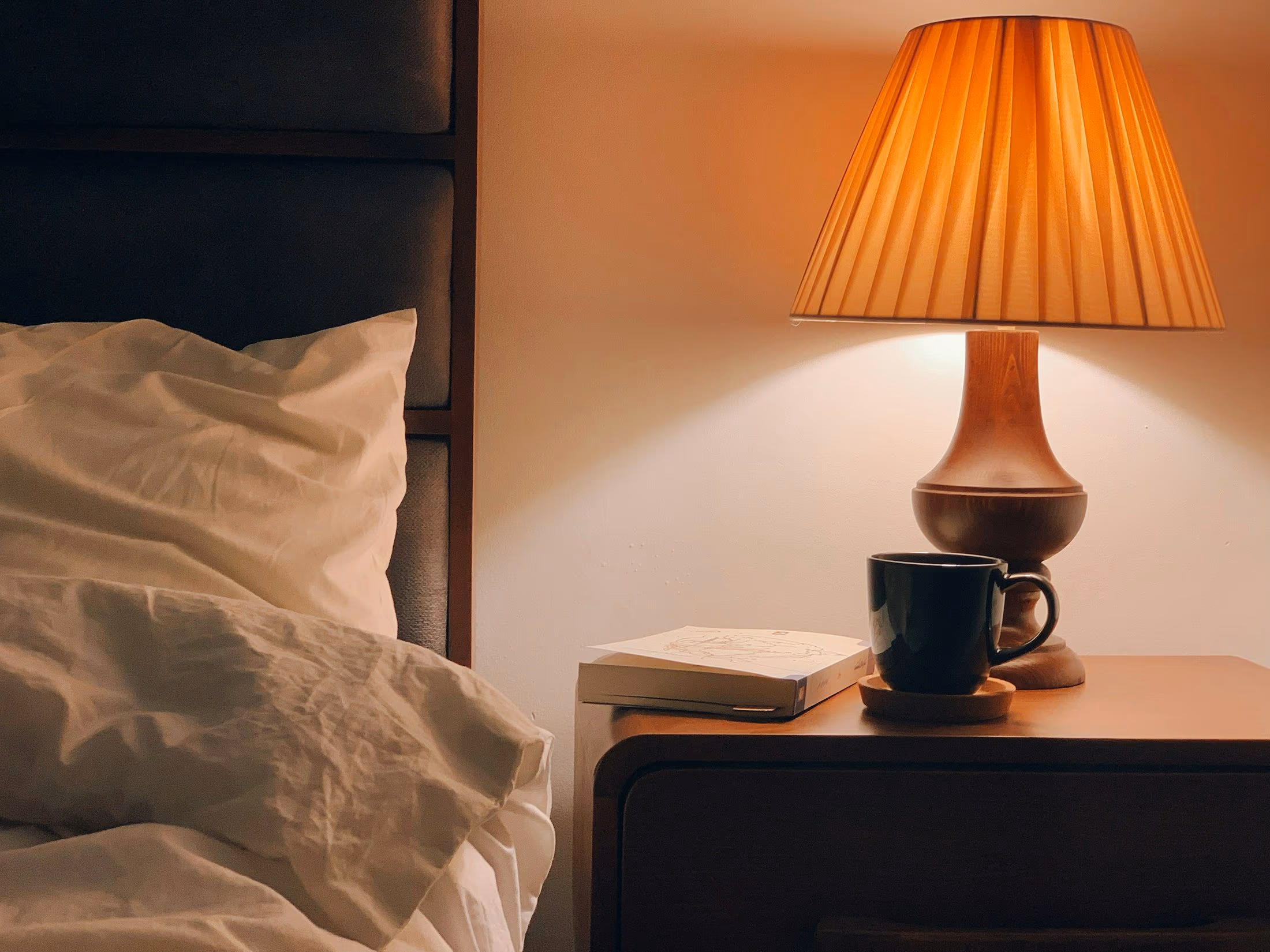 Lampe de chevet à abat-jour plissé allumée sur une table de nuit en bois, près d’une tasse sombre, d’un livre et d’un oreiller froissé