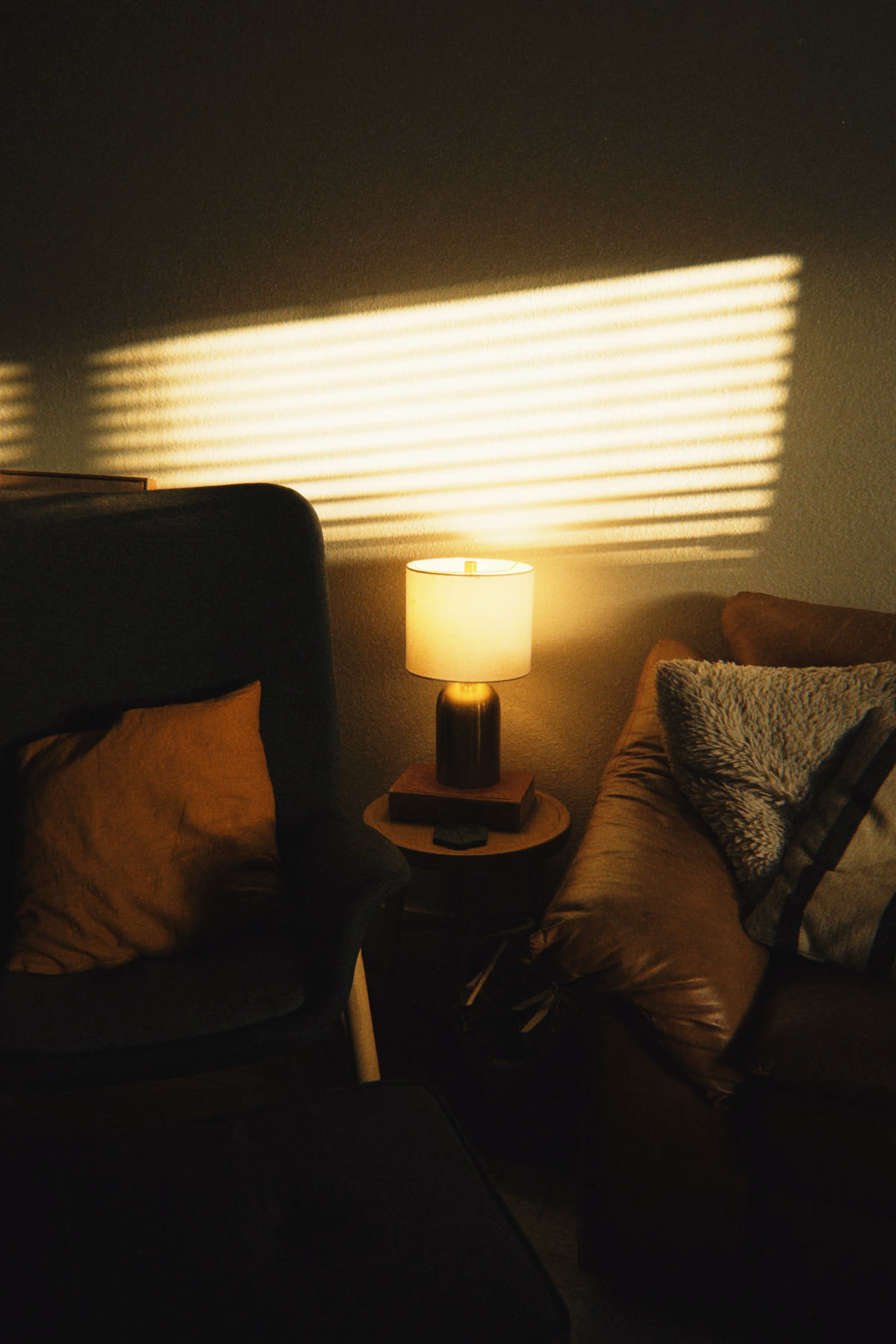 Salon dans la pénombre : lampe à abat-jour allumée sur un guéridon entre un canapé et un fauteuil garnis de coussins