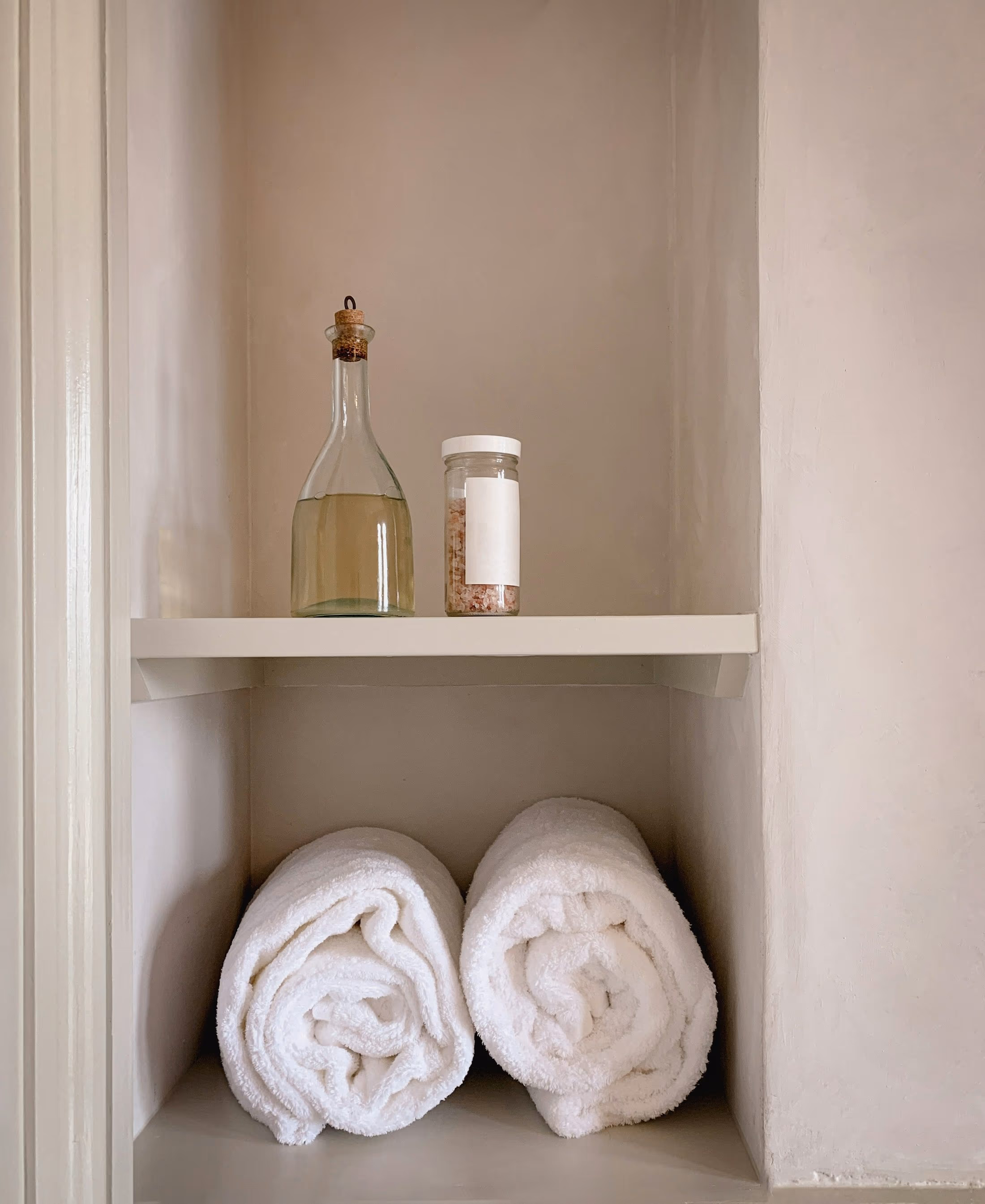 Niche murale crème : carafe en verre bouchée de liège et bocal de cristaux rosés sur l’étagère, deux serviettes blanches roulées en dessous
