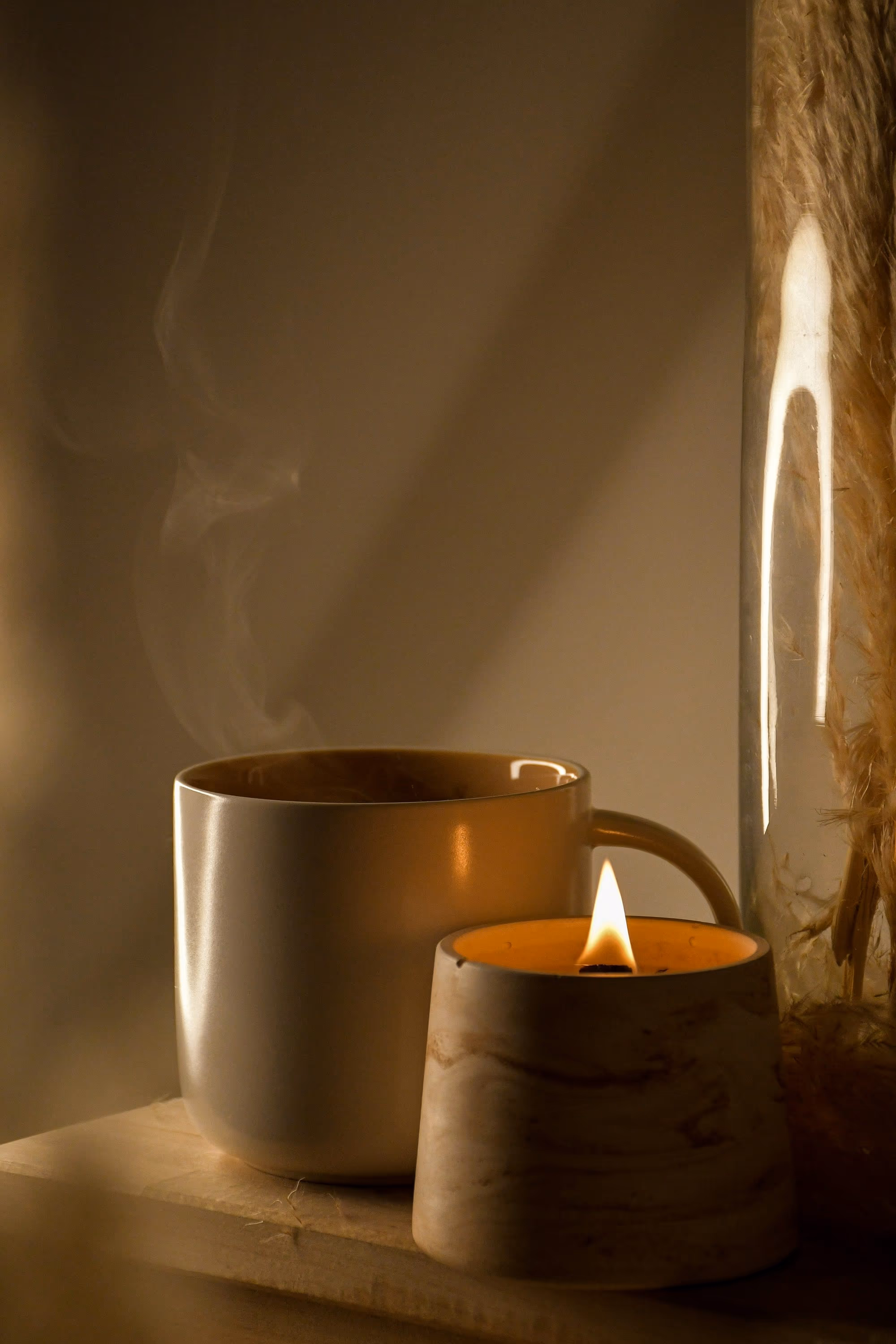 Tasse fumante en céramique crème à côté d’une bougie allumée dans un pot en bois, herbes de la pampa en arrière-plan