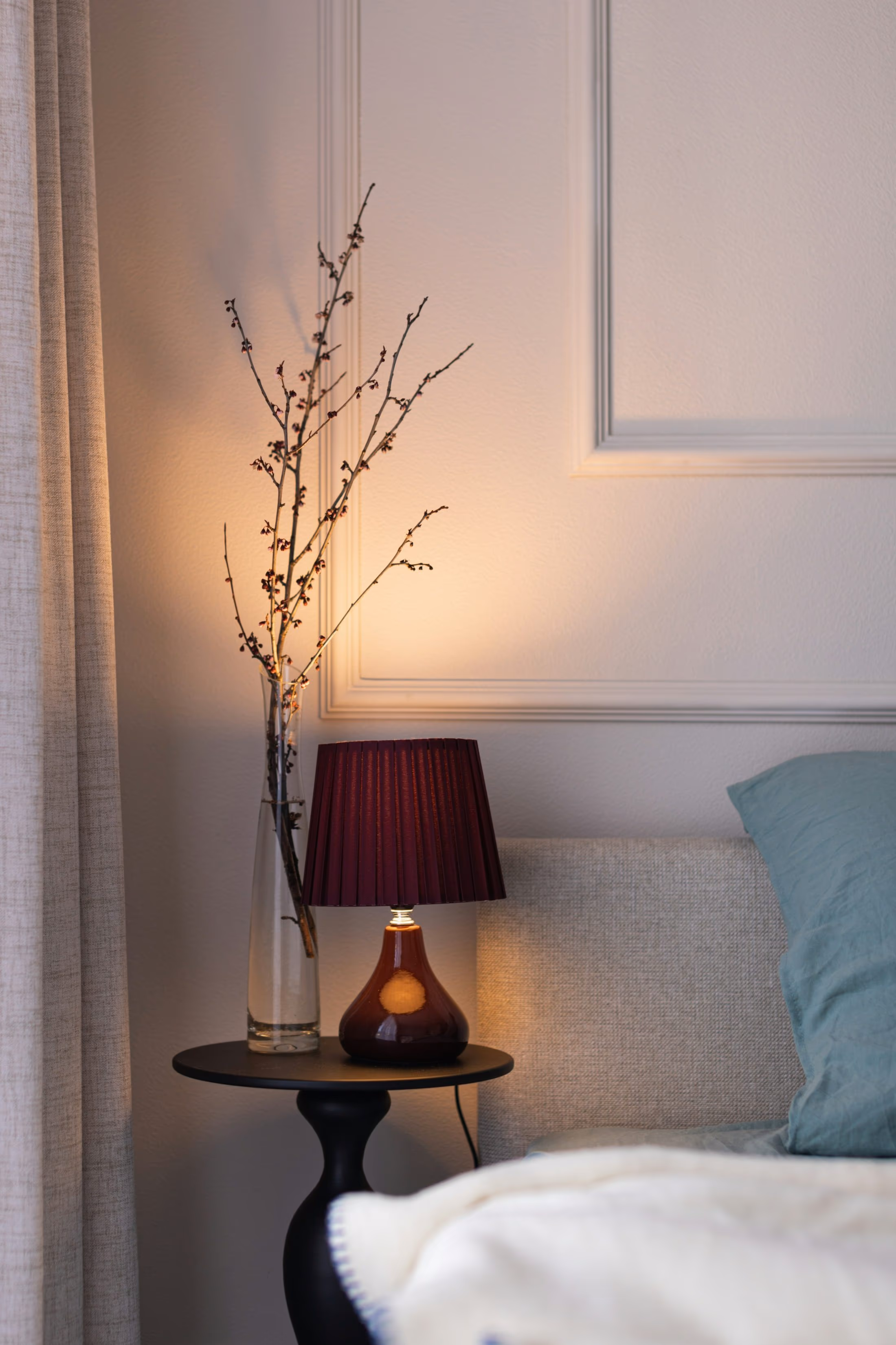 Table de chevet ronde et noire portant une lampe allumée à abat-jour plissé et un vase de branches fines, à côté d’un lit au plaid crème