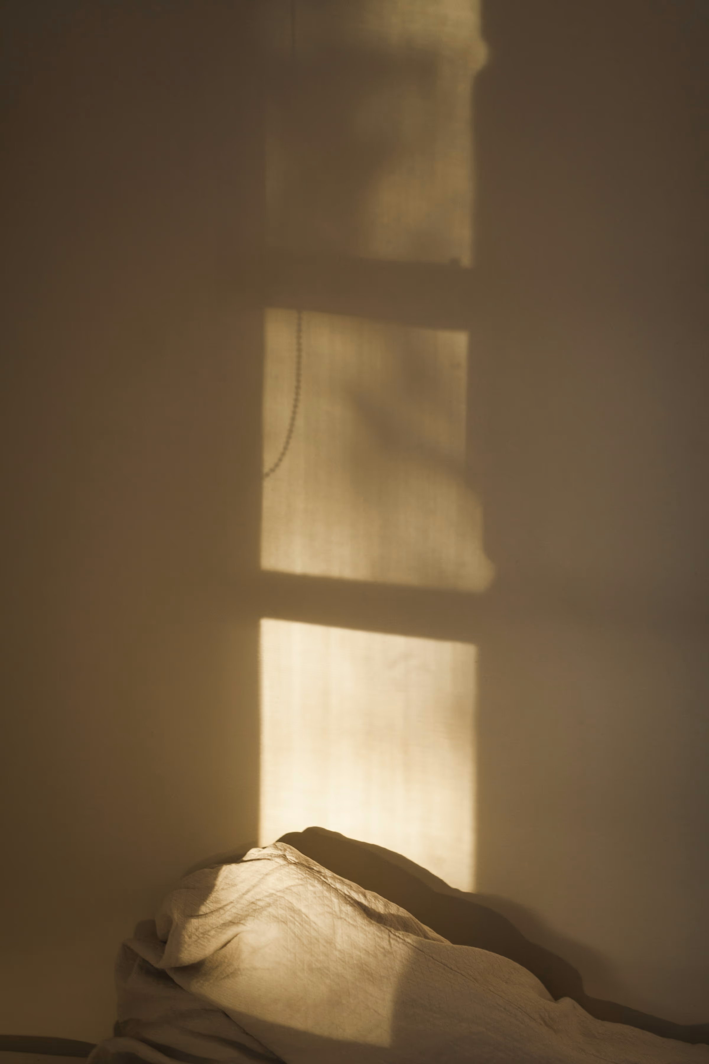 Rai de lumière dorée découpé par une fenêtre sur un mur beige, au-dessus d’un linge de lit froissé dans la pénombre.