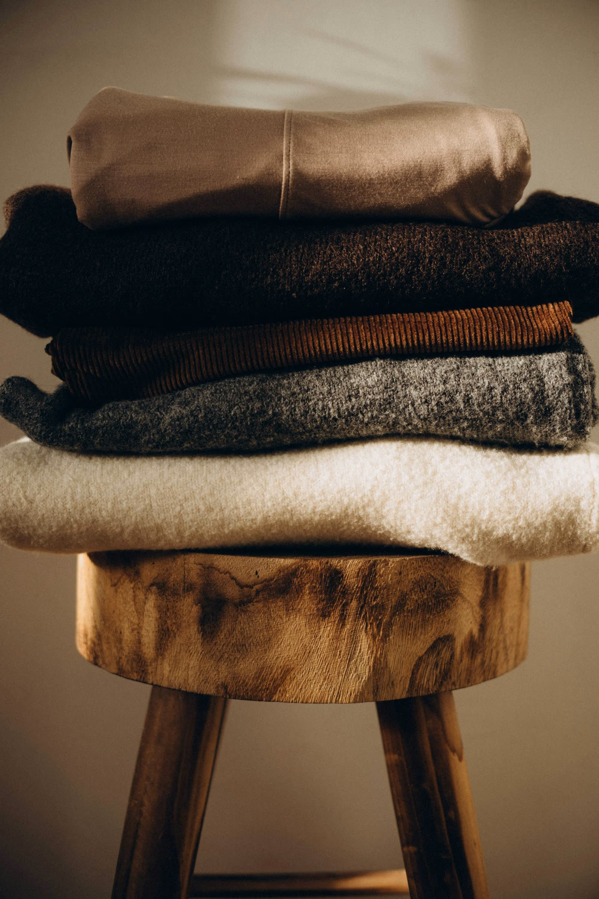 Pile de lainages pliés, du crème au brun, sur un tabouret en bois brut dans une lumière douce