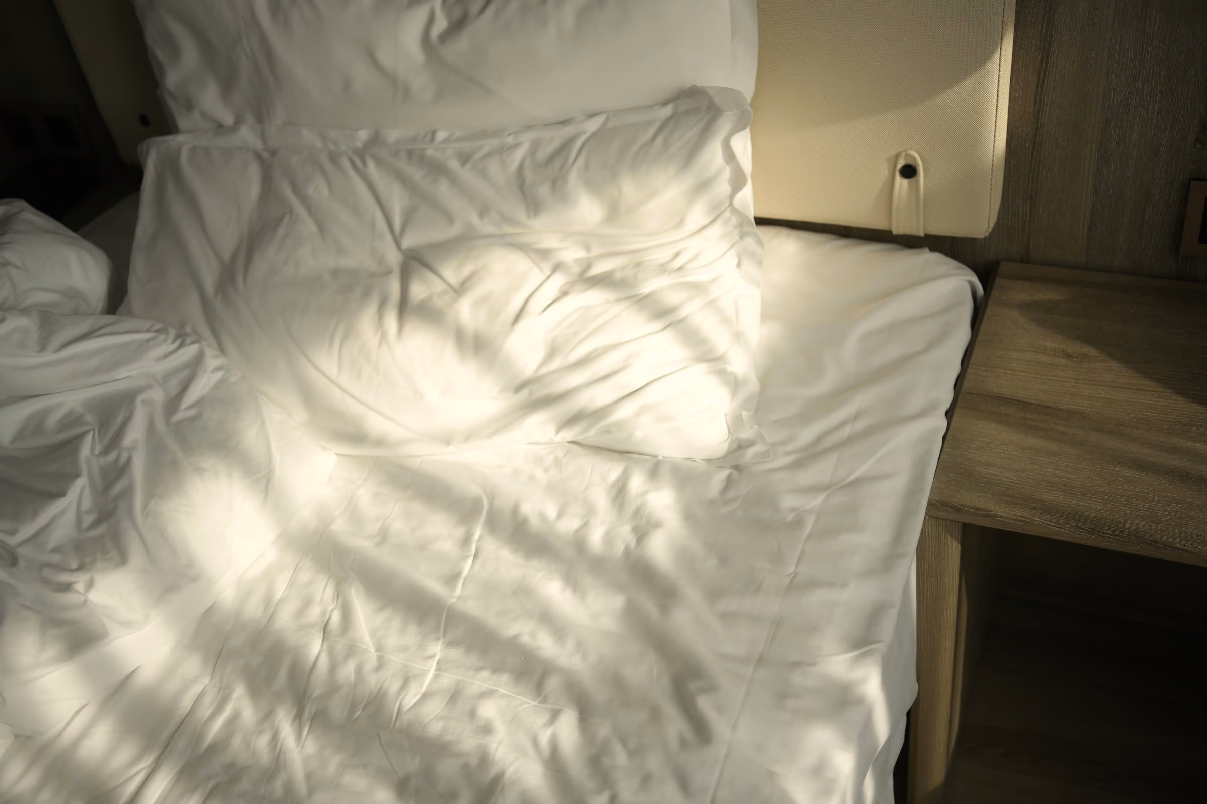 Lit défait aux draps blancs marqués d’ombres de feuillage, à côté d’une table de nuit en bois clair